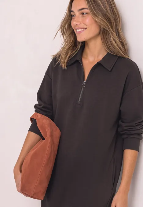 Femme en robe noire à manches longues zippée, souriante, tenant un grand sac marron.