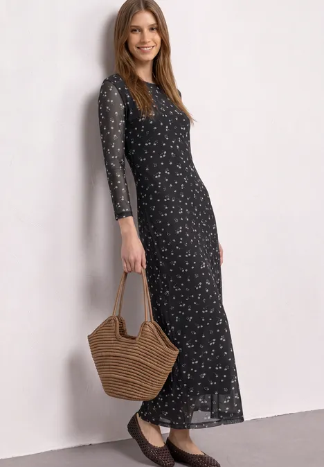 Une femme porte une longue robe noire à motifs floraux et tient un sac en osier.