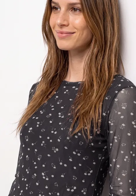 Femme portant un haut noir légèrement transparent orné de petits motifs floraux blancs, avec un sourire discret.