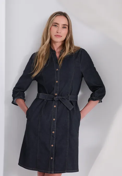 Knielanges Denim Kleid mit Gürtel Rinse Soft Wash Frau in jeansblauem Hemdkleid mit Gürtel, Hände in den Taschen, frontal vor neutralem Hintergrund.
