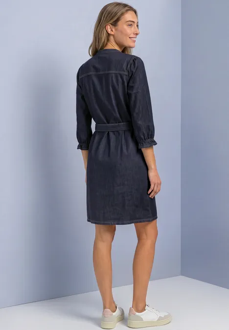 Knielanges Denim Kleid mit Gürtel Rinse Soft Wash Frau von hinten in lässigem dunkelblauen Jeans-Kleid mit 3/4-Ärmeln und Gürtel, dazu helle Sneaker.