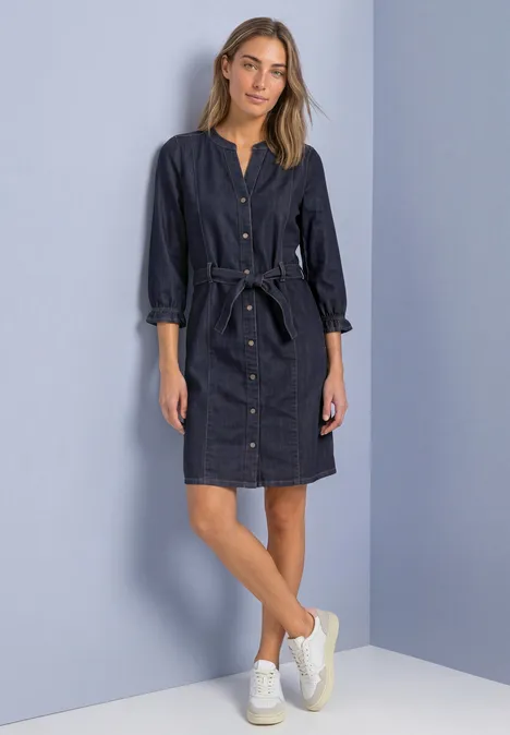 Knielanges Denim Kleid mit Gürtel Rinse Soft Wash Eine Frau trägt ein dunkles Jeans-Hemdkleid mit Gürtel und weißen Sneakers, sie steht vor hellem Hintergrund.