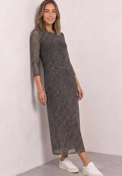 Frau in langem, gemustertem Kleid mit 3/4-Ärmeln und weißen Sneakers vor hellem Hintergrund.