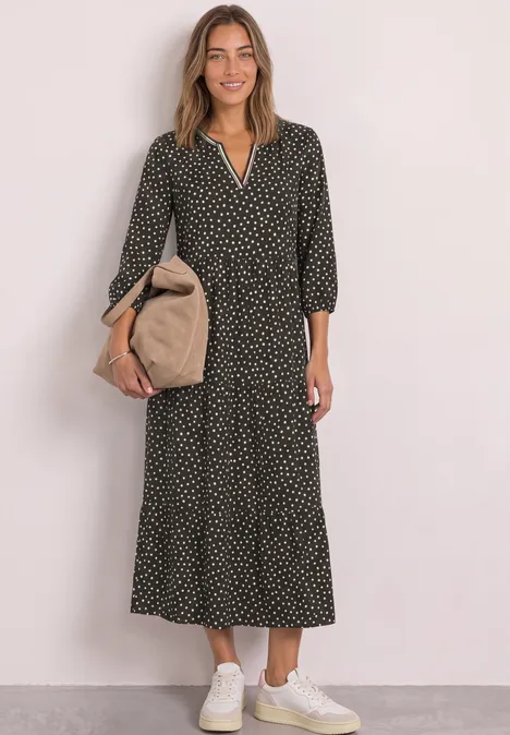 Frau in langem schwarzen Polka-Dot-Kleid mit beigem Beutel und weißen Sneakern vor hellem Hintergrund.