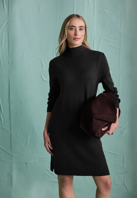 Femme en robe pull noire à col roulé, portant un sac bordeaux devant un fond vert pâle.