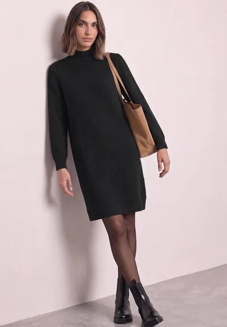 Femme en robe-pull noire, collants et bottines noires, sac bandoulière marron sur fond clair.