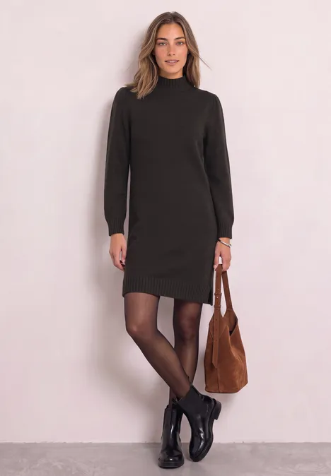 Femme en robe noire à col montant, collants noirs, bottines et sac marron, devant un mur clair.