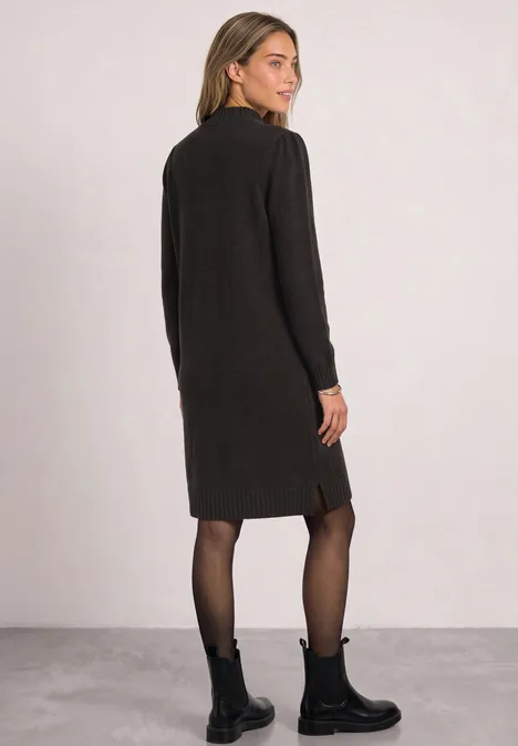 Femme en robe en maille noire mi-longue à manches longues, regard par-dessus l’épaule, boots noires et collants fins.