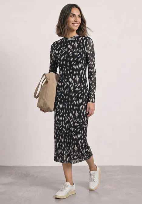 Vrouw draagt zwarte midi-jurk met dessin, witte sneakers en houdt een strooien tas vast.