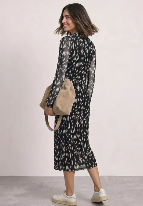 Vrouw in zwarte, dessin midi-jurk met witte sneakers en beige schoudertas tegen neutrale achtergrond.