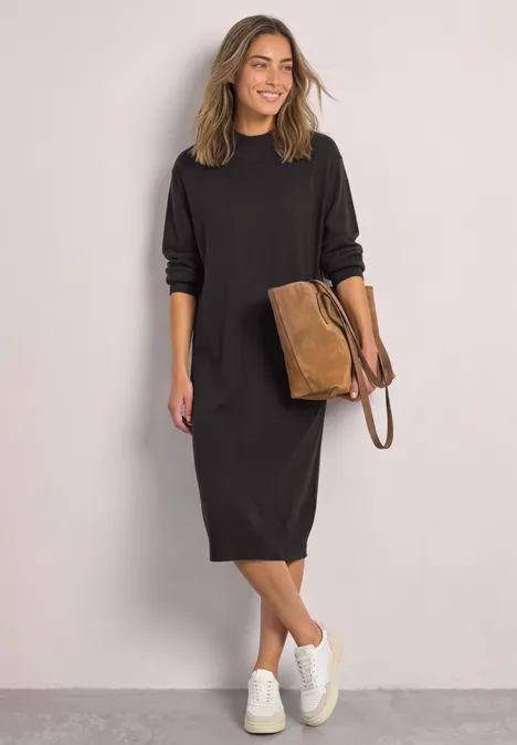 Vrouw in losse zwarte midijurk, witte sneakers en bruine tas, glimlachend voor een grijze achtergrond.