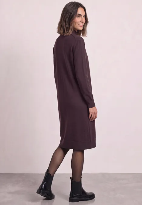 Femme en robe midi violet foncé à manches longues, collants et bottines noires, posée de profil.