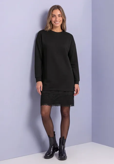 Femme en robe-pull noire à dentelle, collants noirs et bottines devant un mur violet clair.