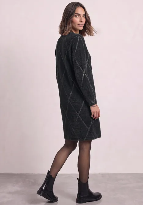 Vrouw in zwarte oversized gebreide jurk met ruitmotief, kijkt over haar schouder, draagt zwarte laarsjes en panty.