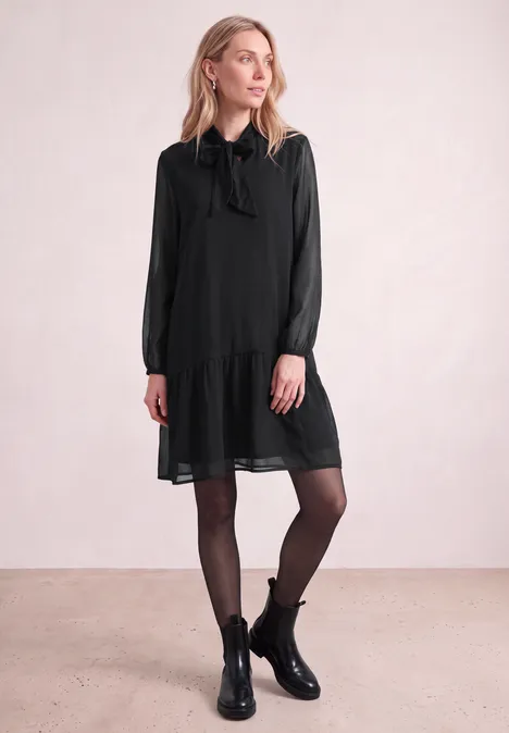 Een vrouw voor een lichte achtergrond draagt een korte zwarte jurk met lange mouwen en strik, zwarte panty's en laarzen.