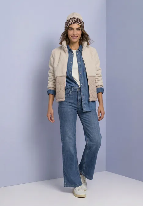 Teddyjas met softshell-details Soft Fawn Beige Frau in Jeansoutfit mit heller Jacke und gemustertem Hut vor violettem Hintergrund.