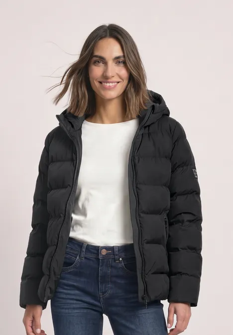 Frau mit schwarzer Steppjacke, Kapuze, weißem Shirt und blauen Jeans lächelt.