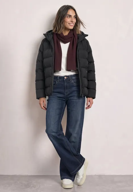 Eine Frau trägt einen schwarzen Steppmantel, blaue Jeans, weiße Sneaker und einen dunklen Schal.