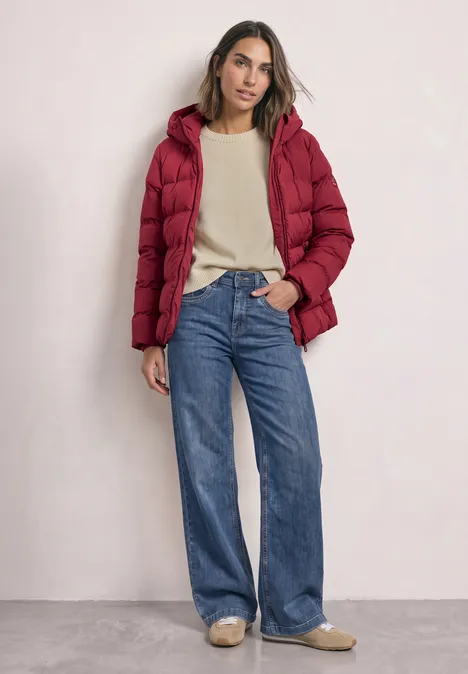 Frau in roter Daunenjacke, beigem Pullover und weiten Jeans vor hellem Hintergrund.
