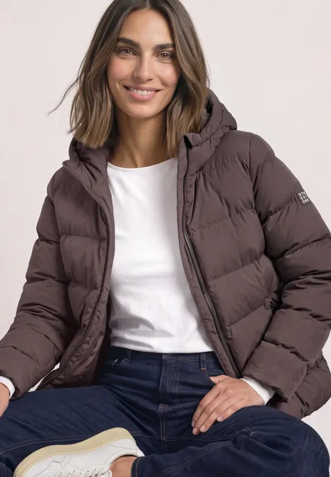 Kurze Jacke mit geklebter Steppung plummy brown Eine lächelnde Frau sitzt und trägt eine schokoladenbraune, gesteppte Winterjacke mit Kapuze.