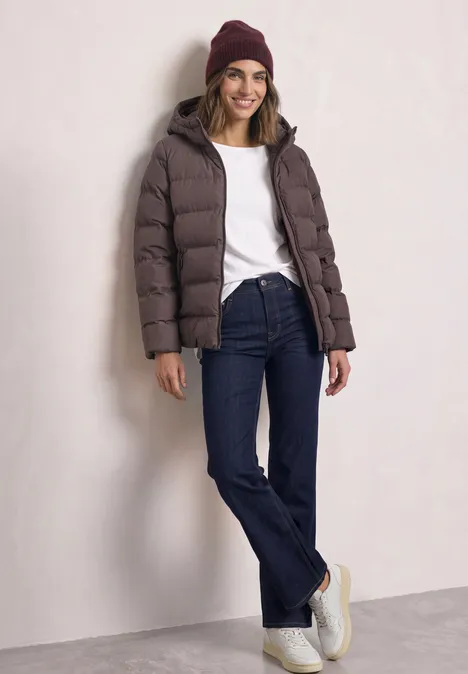 Kurze Jacke mit geklebter Steppung plummy brown Frau mit brauner Steppjacke, dunkelblauen Jeans, weißem Shirt und Beanie lehnt lässig an heller Wand.