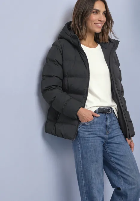 Kurze Jacke mit geklebter Steppung Obsidian Blue Frau in schwarzer Steppjacke, weißem Pullover und blauen Jeans lehnt an Wand und lächelt.