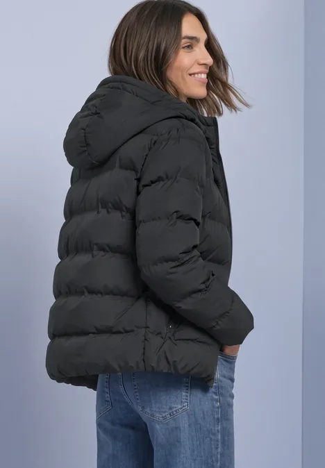Kurze Jacke mit geklebter Steppung Obsidian Blue Frau im Profil trägt eine schwarze Steppjacke mit Kapuze, Hände in den Taschen.