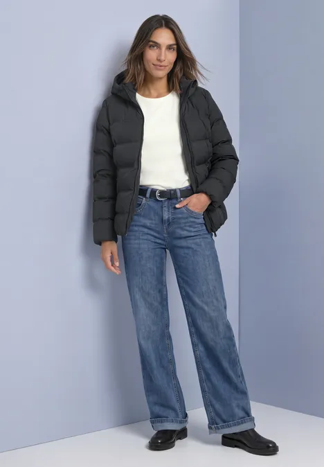 Kurze Jacke mit geklebter Steppung Obsidian Blue Frau in schwarzer Steppjacke, weißem Shirt, blauen Jeans und schwarzen Schuhen vor hellblauem Hintergrund.