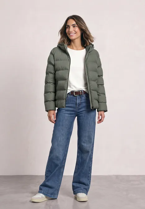 Kurze Jacke mit geklebter Steppung Somber Green Frau in grüner Steppjacke, weißem Shirt und blauen Jeans steht lächelnd vor neutralem Hintergrund.