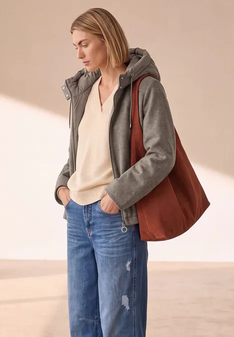 Jacke mit Wollanteil und abnehmbarer Kapuze Muddy Mocha Melange Frau in grauer Kapuzenjacke, weißer Bluse, blauen Jeans und brauner Schultertasche, stehend vor hellem Hintergrund.