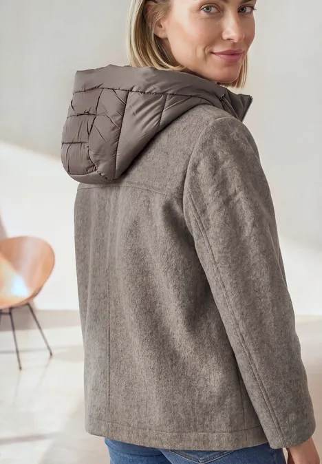 Jacke mit Wollanteil und abnehmbarer Kapuze Muddy Mocha Melange Frau trägt graubraune Jacke mit Kapuze, schaut über die Schulter in minimalistischer Wohnumgebung.