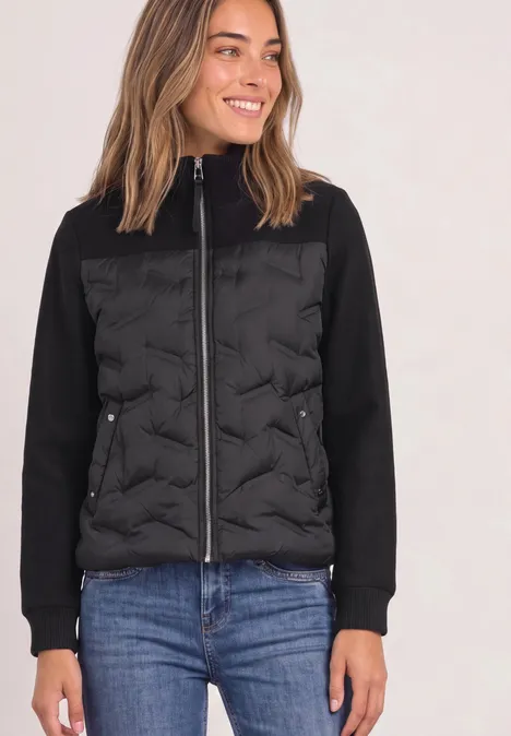 Frau in schwarzer, gesteppter Jacke mit Reißverschluss und blauen Jeans, lächelnd.