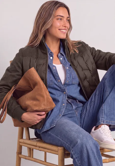 Jonge vrouw in denim top en broek met groene doorgestikte jas, witte sneakers, zittend op kruk, houdt een bruine tas.