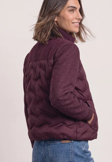 Femme aux cheveux mi-longs porte une veste matelassée bordeaux et un jean, souriante de profil.