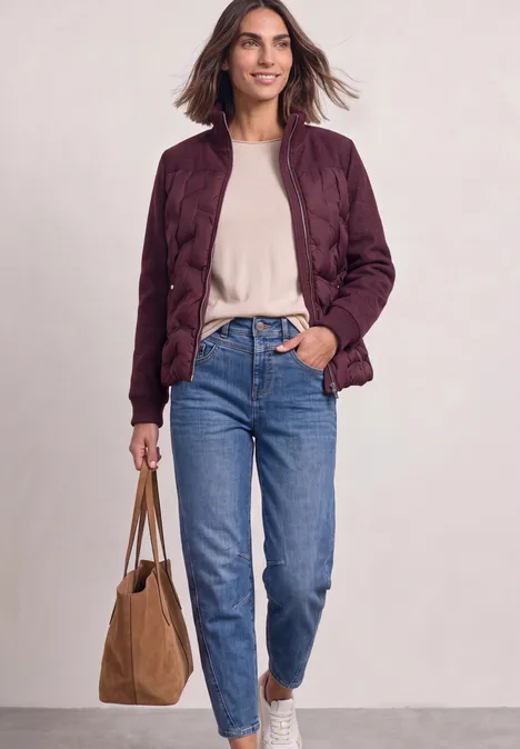 Femme avec une veste matelassée bordeaux, un haut beige, un jean bleu et un sac marron.