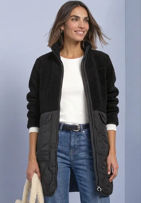 Mantel mit Strukturmix und Stehkragen Black Frau in schwarzer Fleecejacke mit Steppdetails, weißem Shirt und Jeans, hält eine helle Tasche.