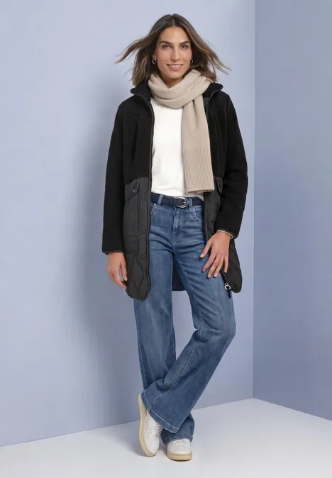 Mantel mit Strukturmix und Stehkragen Black Eine Frau in Jeans, hellem Pullover, Schal und schwarzer Jacke lächelt vor hellblauem Hintergrund.