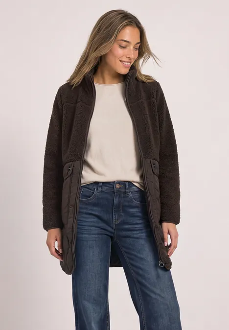 Vrouw in bruine jas, wit shirt en spijkerbroek, glimlachend, kijkt naar links.