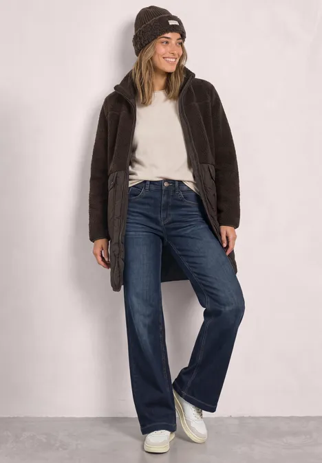 Vrouw lacht in gebreide muts, wit shirt, bruine jas, rechte jeans en witte sneakers voor neutrale achtergrond.