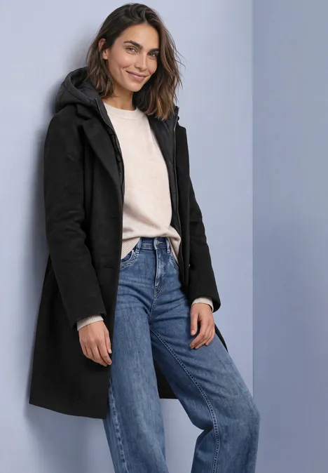 Jas met reverskraag en capuchoninzetstuk Black Frau in einem schwarzen Mantot, hellem Pullover und blauen Jeans, lächelnd vor neutralem Hintergrund.