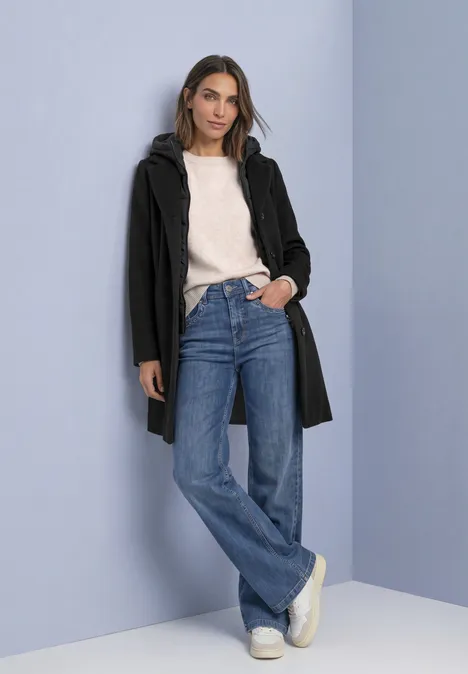 Jas met reverskraag en capuchoninzetstuk Black Frau lehnt locker an hellblauer Wand, trägt schwarzen Mantel, weißen Pullover, blaue Jeans und weiße Sneakers.