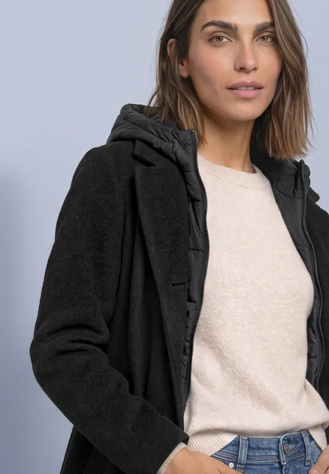 Jas met reverskraag en capuchoninzetstuk Black Frau mit schwarzem Kapuzenmantel, wattiertem Innenfutter, beigem Pullover und Jeans vor schlichtem Hintergrund.