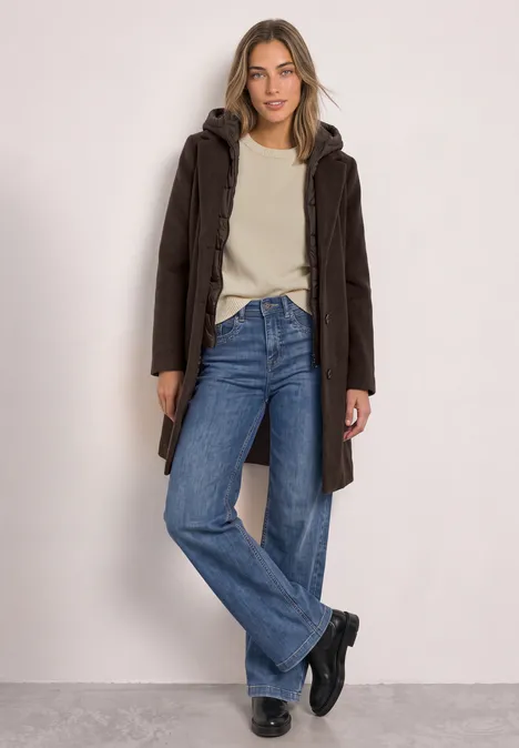 Frau trägt dunklen Mantel mit Kapuze, beigen Pullover, weite Jeans und schwarze Stiefeletten vor hellem Hintergrund.