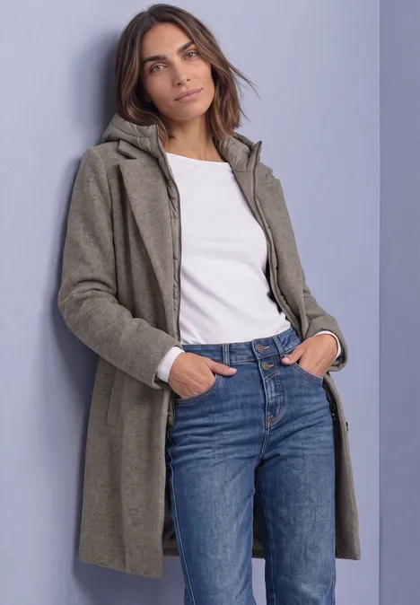 Frau in grünlich-grauem Mantel, weißem Shirt und Jeans lehnt lässig an Wand.