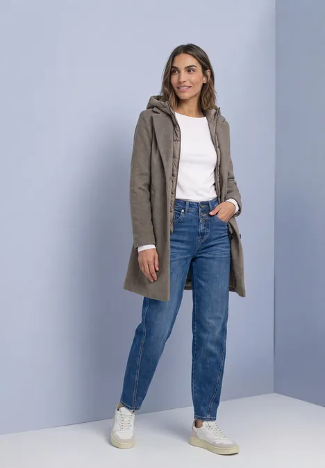 Frau trägt khaki Mantel mit Kapuze, weiße Bluse, blaue Jeans und weiße Sneaker.