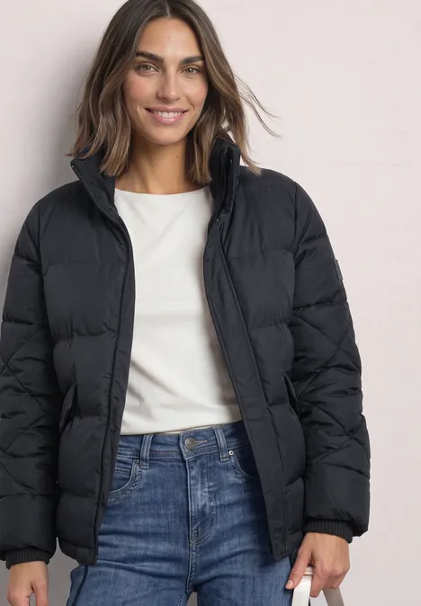 Frau in schwarzer Steppjacke, weißem Shirt und Jeans steht lächelnd vor neutralem Hintergrund.