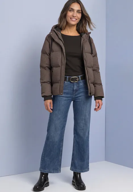 Jacke mit Kapuze und Steppmuster auburn brown Eine Frau posiert lächelnd in einer Daunenjacke, schwarzem Top, blauen Jeans und schwarzen Stiefeletten.