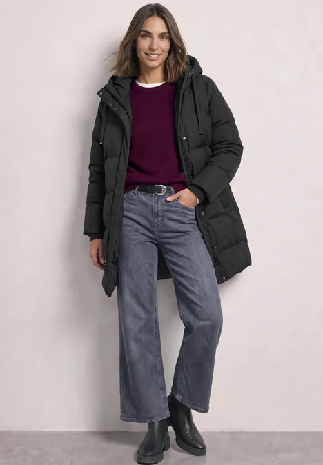 Een lachende vrouw met zwarte gewatteerde jas, bordeauxrode trui, wijde jeans en zwarte laarzen.