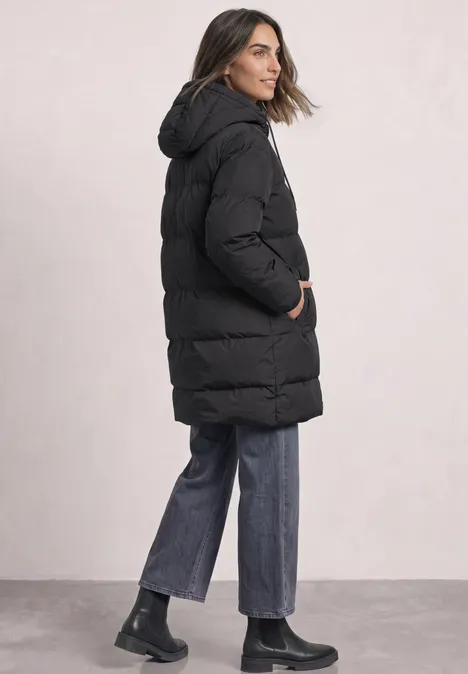 Vrouw in zijaanzicht met zwarte gewatteerde jas met capuchon, blauwe wijde jeans en zwarte boots.