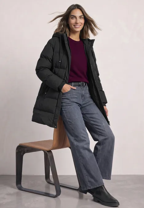 Lachende vrouw in zwarte donzen jas, bordeaux trui, wijde jeans en zwarte laarsjes leunt tegen een kruk.